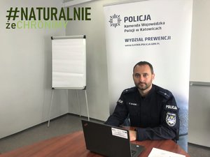 Umundurowany policjant wydziału prewencji siedzi przed laptopem. W tle baner z logo Policji i napisem Komenda Wojewódzka Policji w Katowicach, Wydział Prewencji oraz adresem strony internetowej śląskiej Policji.