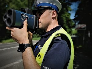 Umundurowany policjant dokonuje pomiaru prędkości. Trzyma laserowy miernik prędkości. W tle znak informujący o końcu obszaru zabudowanego.